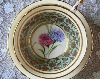 May include: A porcelain teacup featuring a central illustration of pink and blue cornflowers. The interior rim is decorated with a pale green band, intricate gold filigree floral patterns, and a gold-trimmed edge. The handle is white with gold accents.