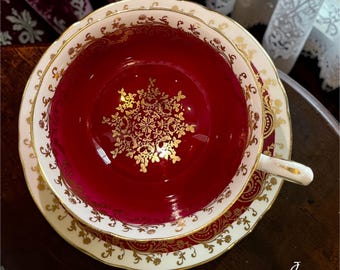 May include: A porcelain teacup and saucer set featuring a deep crimson red interior and rim. The cup and saucer are decorated with intricate gold filigree patterns, floral motifs, and a central gold medallion design inside the cup base.