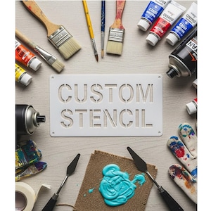 May include: A white stencil with the words "CUSTOM STENCIL" is surrounded by art supplies. Paintbrushes, paint tubes, spray paint, palette knives, and a blob of turquoise paint are arranged on a light wood surface. Paint-covered gloves and tape are also visible.