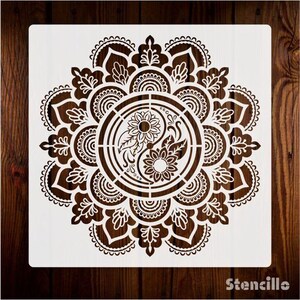 May include: White stencil with a mandala design featuring a yin yang symbol with sunflowers inside. The stencil is on a brown wooden background.