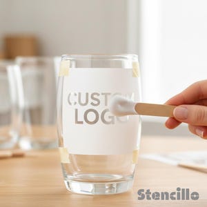 Peut inclure: Un verre transparent avec les mots "CUSTOM LOGO" pochoirés sur une étiquette blanche. Un bâton en bois avec de la peinture blanche est utilisé pour appliquer la peinture. Le mot "Stencillo" est en bas.