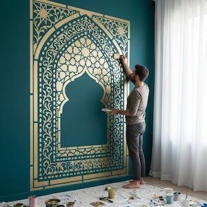 Può includere: Un grande stencil da parete dorato con un intricato design ad arco, motivi geometrici e floreali applicato su una parete color verde ottanio scuro. Lo stencil crea una cornice decorativa con stelle e volute che copre gran parte della superficie della parete.