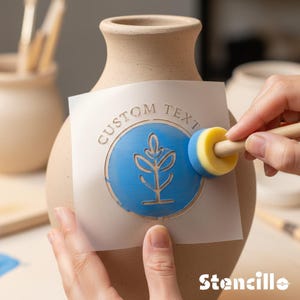 Könnte beinhalten: Eine beige Keramikvase mit einem blau-weißen Schablonenmuster. Die Schablone zeigt ein Pflanzenmotiv und die Worte "CUSTOM TEXT" in einem Kreis. Ein Schwammbürste wird zum Auftragen der Farbe verwendet. Das Wort "Stencillo" steht unten.