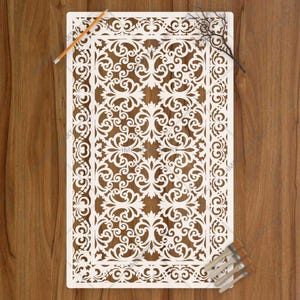 May include: A rectangular white stencil featuring an intricate, symmetrical damask pattern with ornate scrollwork and floral motifs. The stencil is positioned alongside a thin orange pencil and a small cluster of paintbrushes with wooden handles.