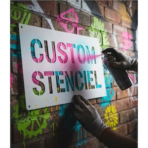 Può includere: Uno stencil bianco con le parole "CUSTOM STENCIL" ritagliate, tenuto contro un muro di mattoni. Lo stencil viene spruzzato con vernice rosa e blu, creando un colorato effetto graffiti. Il muro di mattoni presenta ulteriori graffiti colorati.