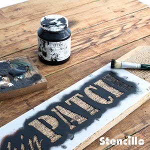 May include: A black jar of Fusion Mineral Paint is on a wooden surface beside a wooden sign stencilled with the word "PATCH" in black. A paintbrush and a small wooden palette with black paint are also visible.