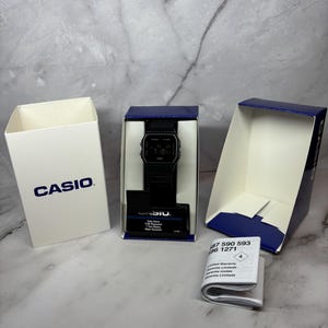 May include: A black Casio digital watch with a black band in its original box. The watch face displays the time, date, and day of the week. The box is white with the Casio logo printed on the front.