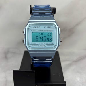 May include: A light blue Casio F-91W digital watch with a black band. The watch face displays the time, date, and day of the week. The watch is water resistant and has a daily alarm and stopwatch.