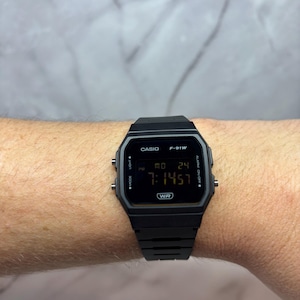 May include: A black Casio F-91W digital watch with a black band. The watch face displays the time, date, and day of the week. The watch is water resistant.