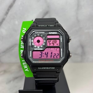 May include: Black Casio digital watch with a pink face and a black resin band. The watch has a world time function, a 10-year battery, and an illuminator. The watch is displayed on a black box with the word "ASIO" printed on the front.