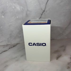 May include: A white box with the word "CASIO" printed in blue on the front. The box has a blue and yellow label on the top.