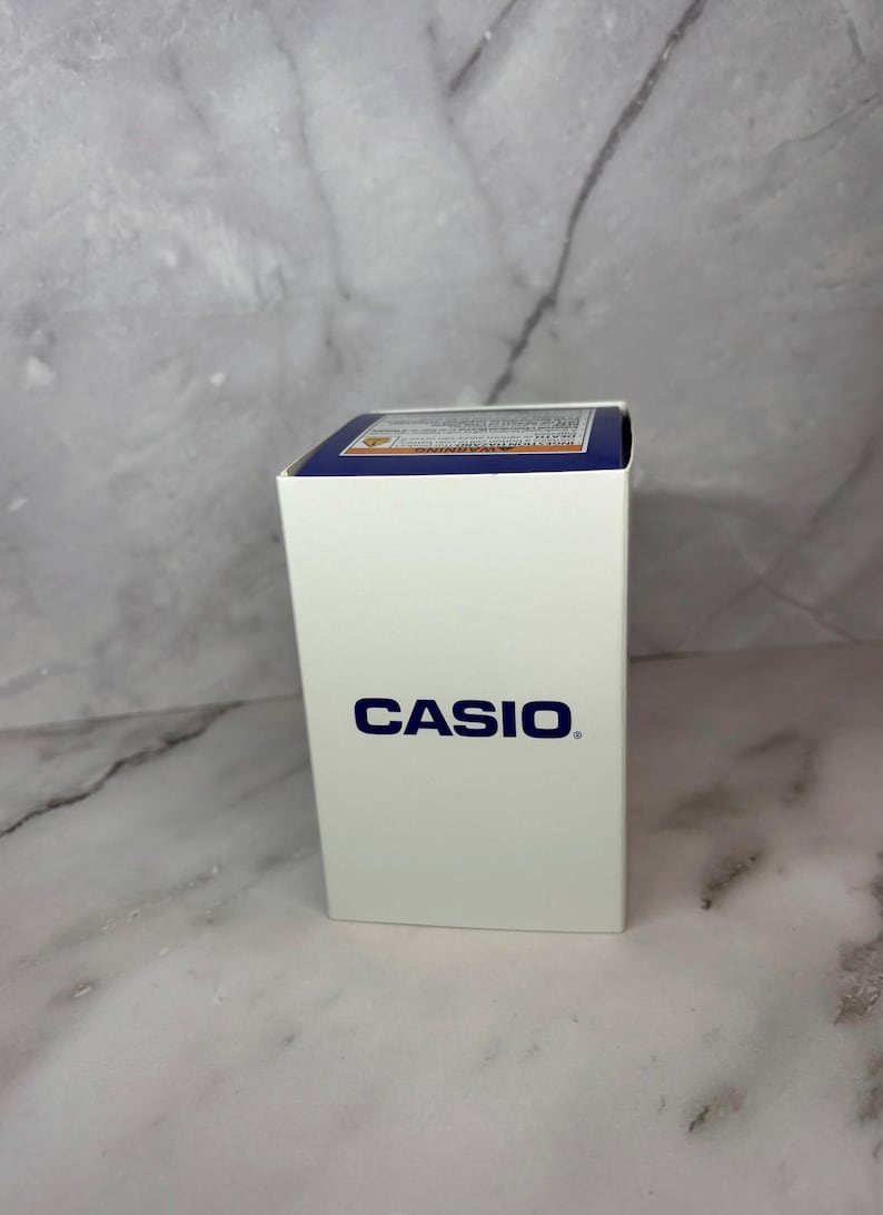 May include: A white box with the word "CASIO" printed in blue on the front. The box has a blue and yellow label on the top.
