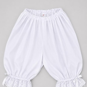 May include: White bloomers with an elasticated waistband and lace trim at the cuffs. The bloomers have a gathered, voluminous shape and are tied with small white bows at the ankles. The fabric appears to be a lightweight cotton or linen.