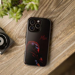 May include: Black phone case featuring a racing helmet design with the name "Max" in red script. The case has a glossy finish and a camera cutout. The phone case is on a wooden surface.