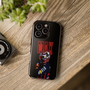 May include: Black phone case featuring a Formula 1 driver in a racing suit and helmet. The word "MAX" is displayed in red neon-style lettering. The case is on a wooden surface.