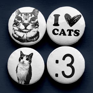 May include: Four white round buttons with black designs. Two buttons feature cat illustrations, one says "I <3 CATS", and the last one has a smiley face with a heart symbol. The buttons are a cat-themed accessory.