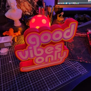 May include: A pink and yellow neon sign with the words "good vibes only" in a retro font. The sign is lit up and has a 3D effect.