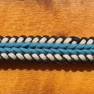 Puede incluir: Una correa trenzada con una sección central azul claro flanqueada por secciones blancas y negras. La correa tiene cierres metálicos dorados en cada extremo. Este artículo se utiliza probablemente para accesorios para mascotas o aplicaciones similares.