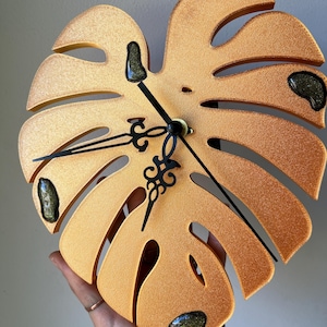 May include: A wall clock shaped like a monstera leaf with a metallic gold finish and a textured surface. The clock features black ornate hands and three small, dark, glittery resin accents embedded within the leaf-shaped frame.