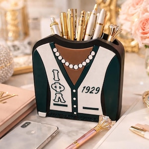 May include: A decorative pen holder shaped like a jacket, in dark green, white, and brown, with the letters "I Φ A" and the number "1929". The holder is filled with gold and white pens. A pink notebook, phone, and watch are also visible.
