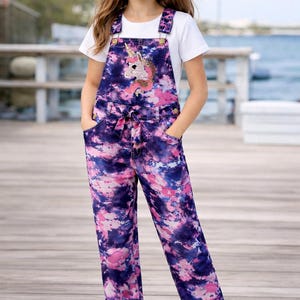 May include: A young person wearing a white t-shirt and a pair of purple, pink, and blue tie-dye overalls with a sequined unicorn design on the bib. The overalls have a matching tie belt and pockets. The person is also wearing a tan baseball cap.