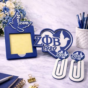 May include: A collection of blue and white Zeta Phi Beta sorority themed desk accessories. Includes a picture frame, heart-shaped decoration, paper clips, and binder clips. The items feature the sorority's logo and a dove symbol.