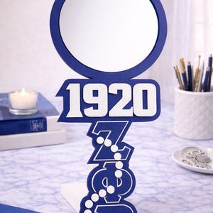 May include: A navy blue and white decorative mirror with a round mirror and a stylized base. The base features the year "1920" and Greek letters. The mirror is on a marble-patterned surface with other decorative items.