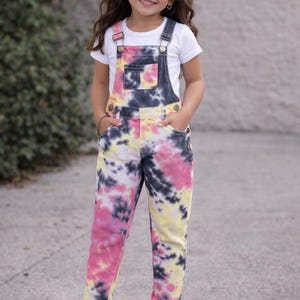 May include: A young girl wearing a white t-shirt and tie-dye denim overalls. The overalls feature a colorful pattern of pink, yellow, and black. She is also wearing white sneakers and a pink bow in her hair.