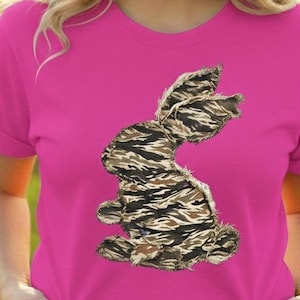 Puede incluir: Una mujer con una camiseta rosa brillante con un diseño decorativo de conejito. El conejito está hecho de un patrón de camuflaje en tonos marrones, beige y negro. La camiseta es de color sólido y tiene mangas cortas.
