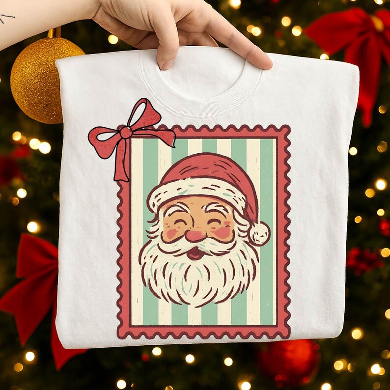 May include: White crew neck t-shirt featuring a Santa Claus graphic. The Santa Claus image is framed within a red postage stamp design with a red bow. The Santa Claus graphic has a red hat, white beard, and a smiling face.