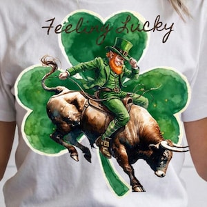 May include: White t-shirt featuring a watercolor illustration of a leprechaun riding a bull in front of a green shamrock. The text "Feeling Lucky" is written above the shamrock in brown script.