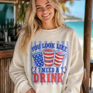 以下が含まれることがあります： 「YOU LOOK LIKE I NEED A DRINK」の文字が青と赤でプリントされたクリーム色のスウェットシャツ。愛国的なデザインの3つの飲み物カップが描かれています。カジュアルで快適なスタイルです。