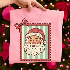 May include: A light pink t-shirt featuring a Santa Claus graphic within a postage stamp design. The Santa illustration has a red hat and a white beard. A red bow is at the top left corner of the stamp.