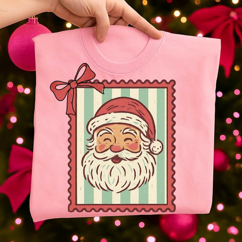 May include: A light pink t-shirt featuring a Santa Claus graphic within a postage stamp design. The Santa illustration has a red hat and a white beard. A red bow is at the top left corner of the stamp.