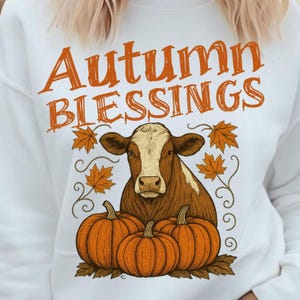 Pode incluir: Camisola branca com o texto "Autumn Blessings" em laranja. O design apresenta uma vaca castanha e branca, três abóboras laranja e folhas de bordo laranja, perfeita para o outono.