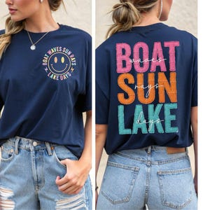 Puede incluir: Camiseta azul marino con un diseño de cara sonriente y las palabras "Boat Waves Sun Rays Lake Days". La parte trasera de la camiseta tiene las palabras "Boat Waves Sun Rays Lake Days" en un diseño apilado. La camiseta se combina con pantalones cortos de mezclilla azul claro.
