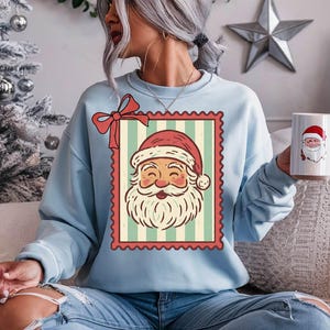 May include: Light blue sweatshirt featuring a vintage Santa Claus graphic within a postage stamp design. The Santa illustration has a red hat and a white beard. A matching mug with Santa's face is also visible.