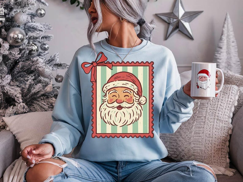 May include: Light blue sweatshirt featuring a vintage Santa Claus graphic within a postage stamp design. The Santa illustration has a red hat and a white beard. A matching mug with Santa's face is also visible.