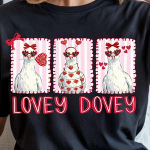 Op de afbeelding: Zwart T-shirt met een ontwerp van drie witte duiven met hartvormige zonnebrillen en strikken. Eén duif houdt een lolly vast. De tekst "LOVEY DOVEY" staat in het rood onder de duiven. Het ontwerp heeft een roze gestreepte achtergrond.
