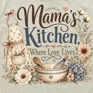 May include: A light gray t-shirt featuring a floral apron design with the words "Mama's Kitchen, Where Love Lives." The design includes a bowl, whisk, and wooden spoon, surrounded by flowers and hearts.