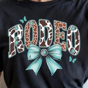 May include: Black t-shirt featuring the word "RODEO" in a western design. The letters have a cowhide and leopard print pattern, with turquoise accents and a large turquoise bow. Two turquoise butterflies are also present.