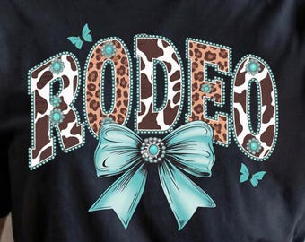 Op de afbeelding: Zwart T-shirt met het woord "RODEO" in een western-stijl ontwerp. De letters hebben een koeienhuid- en luipaardprintpatroon, geaccentueerd met turquoise details en een grote turquoise strik. Twee turquoise vlinders zijn ook aanwezig.