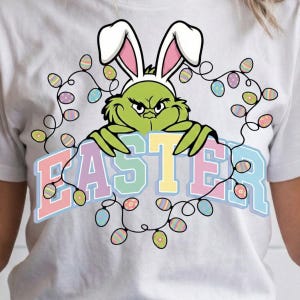 Könnte beinhalten: Weißes T-Shirt mit einer grünen Figur, die Hasenohren trägt und ein Schild mit der Aufschrift "EASTER" in Pastellfarben hält. Das Design beinhaltet eine Lichterkette mit Ostereiern, die einen festlichen und verspielten Look erzeugt.