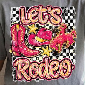 May include: A gray t-shirt featuring a vibrant rodeo-themed graphic. The design includes a pink cowboy boot, a pink cowboy hat with a yellow brim, a pink bucking bronco, and the words "Let's Rodeo" in a playful font. Gold stars and a black and white checkered background complete the design.