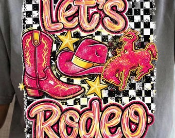 May include: A gray t-shirt featuring a vibrant rodeo-themed graphic. The design includes a pink cowboy boot, a pink cowboy hat with a yellow brim, a pink bucking bronco, and the words "Let's Rodeo" in a playful font. Gold stars and a black and white checkered background complete the design.