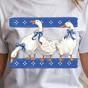 May include: White t-shirt featuring a whimsical illustration of three white geese adorned with blue bows. The geese stand on a blue rectangular banner with white dot accents. The design is centered on the shirt.