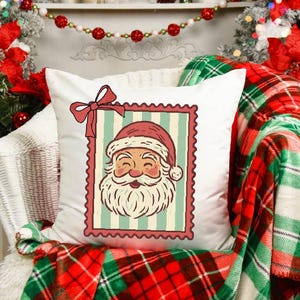 May include: A white square pillow with a Santa Claus design. The Santa image is framed in a postage stamp style with a red border and bow. The pillow is on a chair with a red and green plaid blanket.