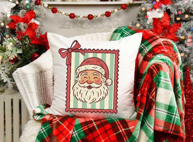 May include: A white square pillow with a Santa Claus design. The Santa image is framed in a postage stamp style with a red border and bow. The pillow is on a chair with a red and green plaid blanket.