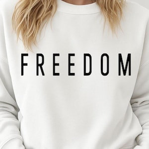 May include: White sweatshirt with the word "FREEDOM" printed in large, bold, black letters across the chest. The sweatshirt is a solid color and has long sleeves. The overall style is casual and comfortable.