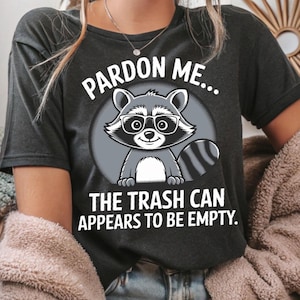 Puede incluir: Camiseta gris oscuro con un mapache de dibujos animados con gafas, dentro de un círculo gris. El texto de arriba dice "Pardon Me..." y debajo, "The Trash Can Appears To Be Empty."
